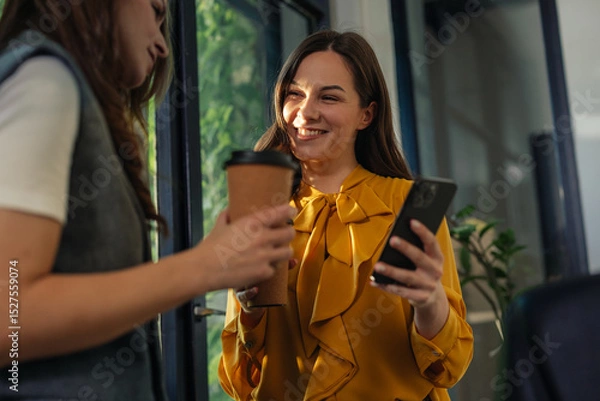 Fototapeta Gen Z businesswomen enjoying coffee break and using smartphone in modern office