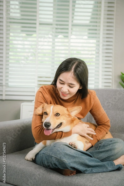 Fototapeta A cheerful young woman sits at home on a comfortable sofa, happily hugging her cute dog in a warm and loving home.