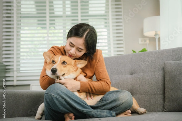 Fototapeta A cheerful young woman sits at home on a comfortable sofa, happily hugging her cute dog in a warm and loving home.