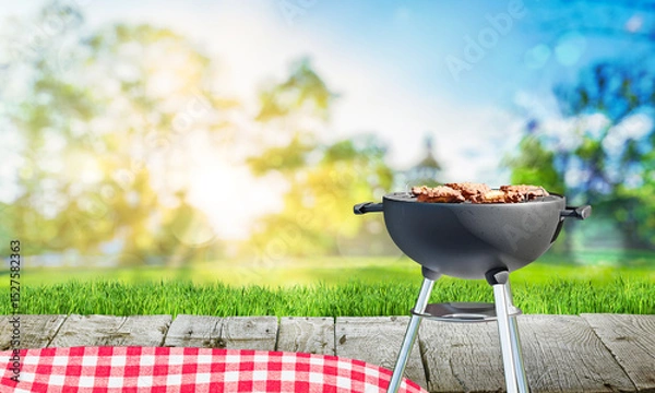 Fototapeta charcoal barbecue grill with open flame, chicken meat, backyard and wooden table with red tablecloth