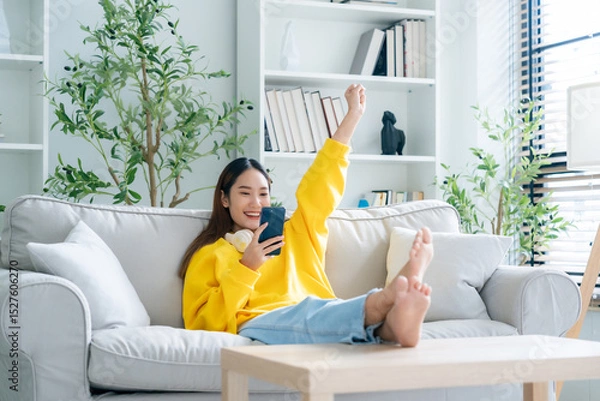 Fototapeta Happy young asian woman relax on comfortable couch at home texting messaging on smartphone, smiling girl use cellphone, chatting online message, shopping online from home