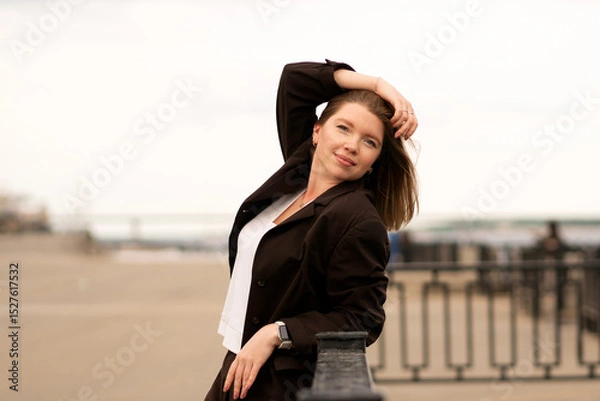 Fototapeta Woman in brown suit resting arm on railing, looking to side outdoors. Natural feel with soft light. Casual, confident pose and expression add genuine and approachable vibe.