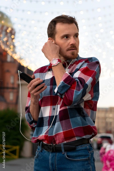 Fototapeta Man adjusting earphones while listening to music under lights. Guy with beard and modern hairstyle wearing in blue, red and white plaid shirt walking through the city streets in urban background