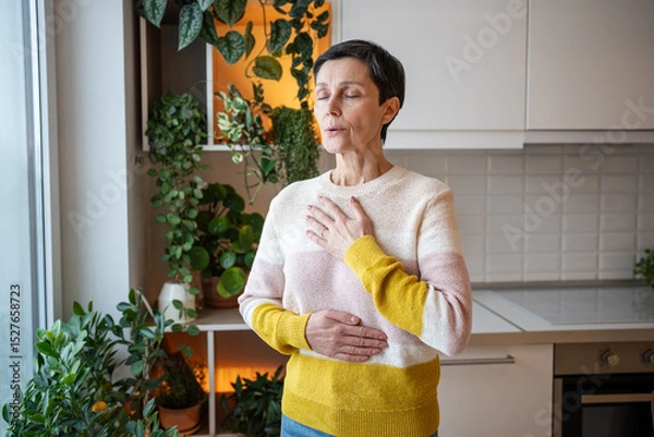 Fototapeta Content serene older woman practicing deep breaths in meditation, hands on torso at home. Stress relief through controlled breathing, comfortable leisure activity, holistic wellness, mindful happiness