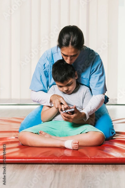 Fototapeta Physiotherapist helping child with cerebral palsy using smartphone during rehabilitation session
