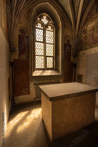Fototapeta Gothic architectural interior features a pointed arch window with ornate tracery and lattice design, allowing softened light into the room. Stone walls are adorned with faded frescoes.