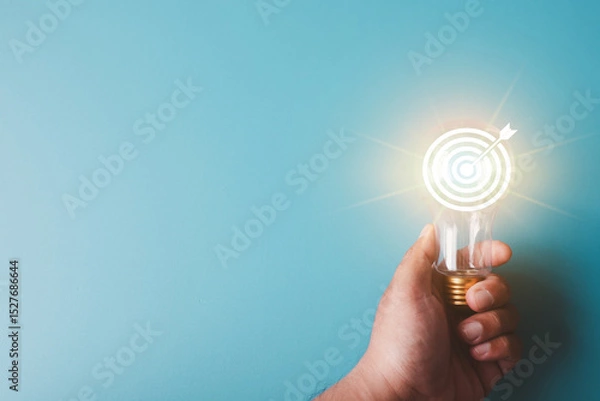 Obraz Hand holding a lightbulb with a glowing target board and arrow icon inside. Concept for business strategy, setting clear objectives, marketing goals, innovative solutions,creative mindset.