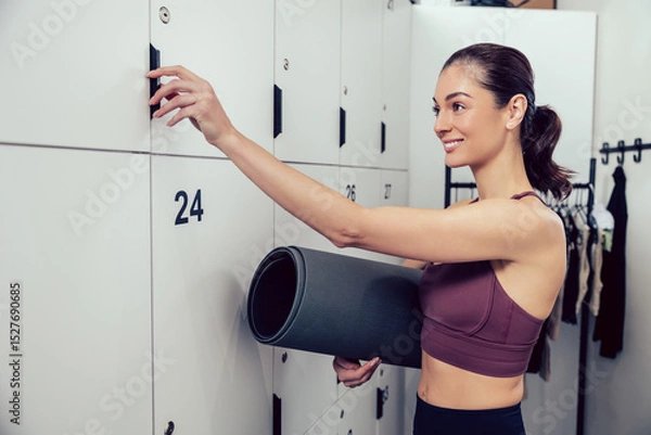 Fototapeta Smiling young woman with a rolled mat opening a locker in the gym