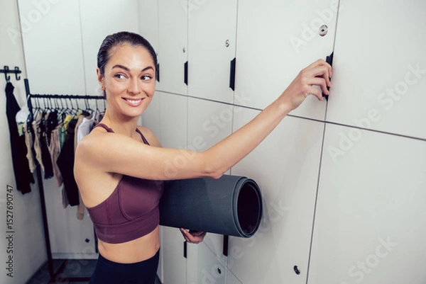 Fototapeta Smiling young woman with a rolled mat opening a locker in the gym