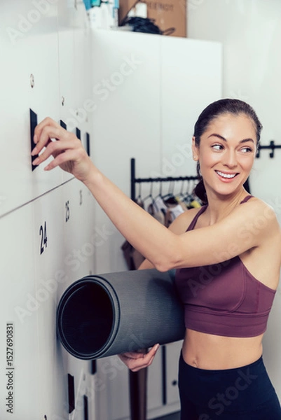Fototapeta Smiling young woman with a rolled mat opening a locker in the gym
