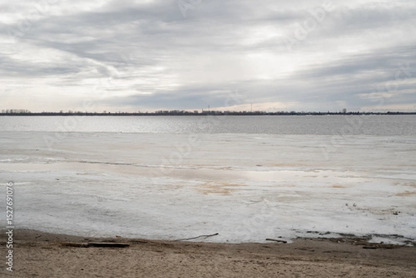 Fototapeta The wide northern river with a sandy bank was partially freed from winter snow and ice on a cloudy day