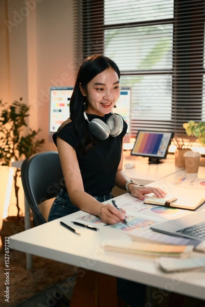 Fototapeta Graphic designer woman is sitting at a desk with a laptop and a notebook. She is wearing headphones and smiling