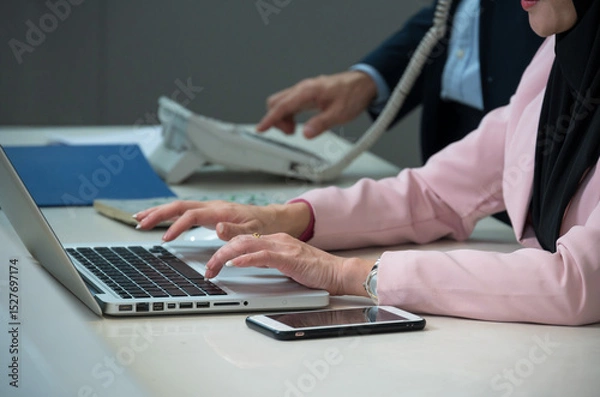 Fototapeta Close up shot of woman in a pink blazer and black hijab typing on  laptop with smartphone on office desk while businessman using landline phone in background