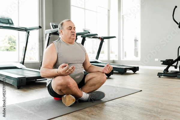 Fototapeta Overweight senior man meditating in a cross-legged pose on a yoga mat at the gym. Promotes mental wellness, mindfulness, and a healthy lifestyle through calmness, balance, and inner peace.