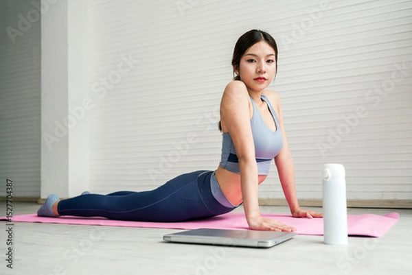 Fototapeta Young woman practices yoga on a pink mat in a bright room, performing cobra pose for flexibility and back strength. Dressed in activewear, she focuses on wellness and balance in a calm setting.