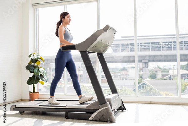 Fototapeta Confident young woman walking on a treadmill in a bright indoor gym with large windows, embracing wellness, fitness, and a healthy lifestyle through daily cardio and mindful exercise routines.
