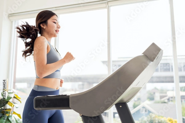 Fototapeta Focused young woman running on a treadmill in a bright modern room, capturing dedication to fitness, cardio training, and healthy lifestyle for wellness, weight loss, and daily exercise routine.