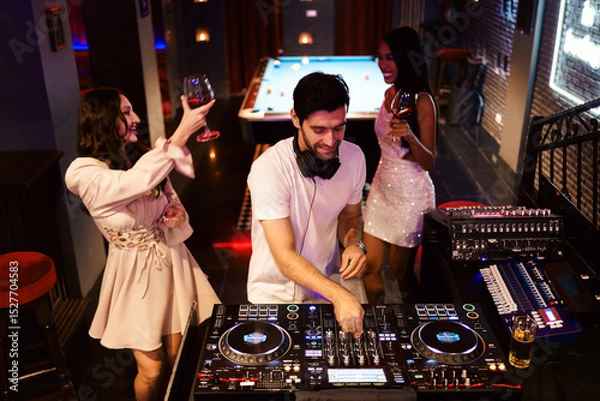 Fototapeta DJ playing lively music in a nightclub as diverse women dance and enjoy drinks together, celebrating nightlife, diversity, and joyful party energy under colorful lights and upbeat vibes.