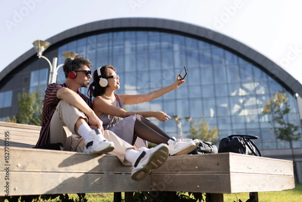Fototapeta Young couple sitting on a bench with headphones, taking a selfie outside modern architectural building on a sunny day