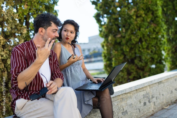 Fototapeta Two people sitting outdoors discussing an idea while using a laptop, showcasing teamwork and communication in a casual setting