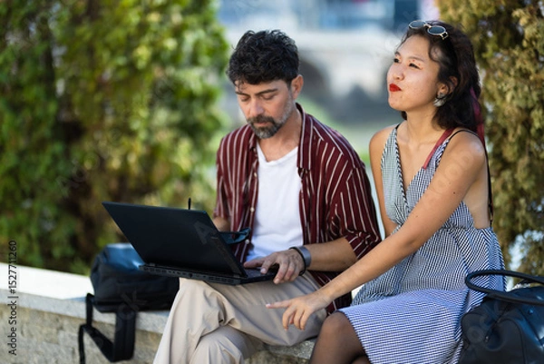 Fototapeta Young professionals collaborating outdoors, working on a laptop together in a sunny park setting, enjoying teamwork and creative brainstorming in a relaxed and natural environment