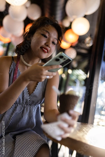 Fototapeta Smiling young woman enjoying ice cream while using smartphone in a lively cafe with glowing lights