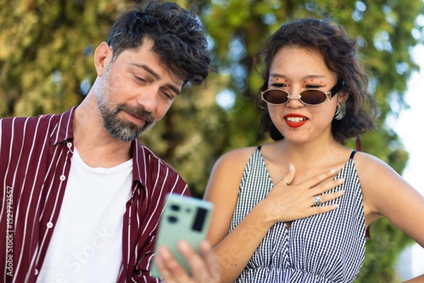 Fototapeta Two friends sharing a moment looking at a smartphone outdoors on a sunny day, showcasing connection and technology in a social setting.