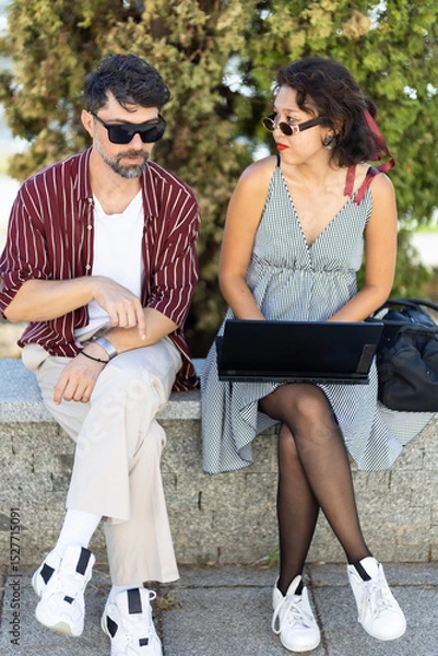 Fototapeta Stylish man and woman in sunglasses engaged in conversation outdoors, sitting on a stone bench with green trees in the background, highlighting casual summer fashion and urban interaction
