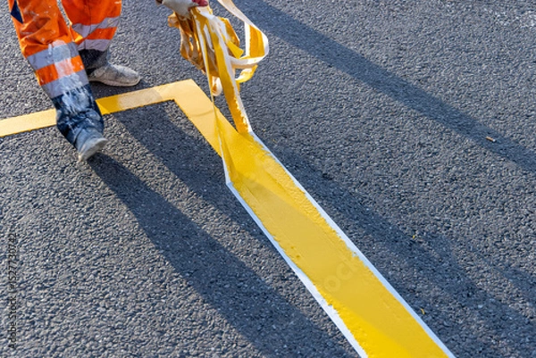 Fototapeta Road worker applying yellow road markings to asphalt using stencil tape. Close-up of a road worker applying yellow paint to asphalt with stencil tape