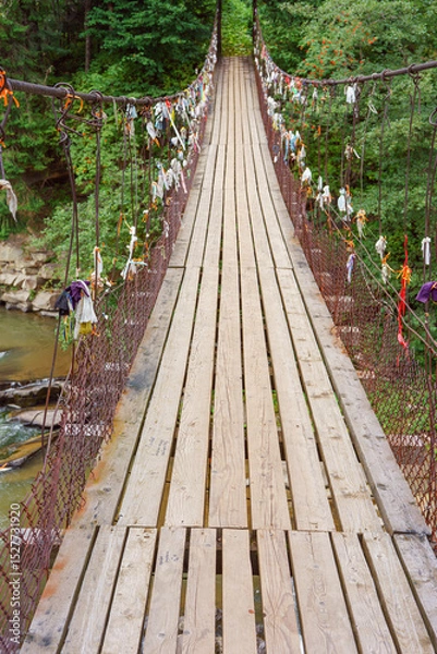 Fototapeta Wooden suspension bridge with colorful ribbons hanging from wires crosses fast mountain river, mountain creek in lush green coniferous Carpathian forest. Adventure, travel, hiking and local charm.