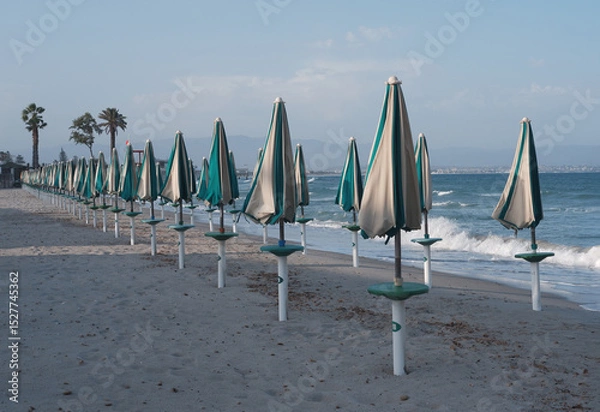 Obraz Closed umbrellas on sandy beach in Sardinia, Poetto shore with summer mood and calm coastal atmosphere