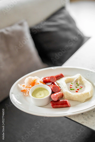 Fototapeta A plated meal featuring sliced sausages, mashed potatoes with garnish, green sauce, and shredded vegetables. The dish is served on a white plate with a blurred background.