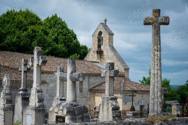 Obraz A cemetery filled with ornate stone crosses stands adjacent to a historic church in Saint-Laurent-des-Combes, surrounded by lush greenery under a dramatic sky.