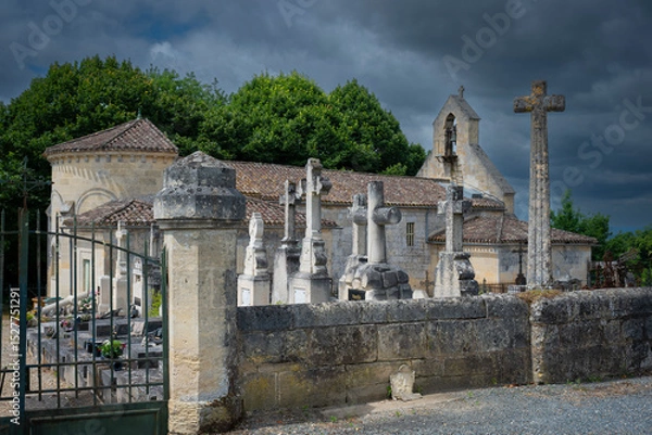 Obraz A cemetery filled with ornate stone crosses stands adjacent to a historic church in Saint-Laurent-des-Combes, surrounded by lush greenery under a dramatic sky.