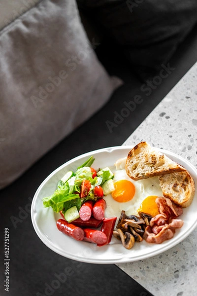 Fototapeta A hearty breakfast plate with sunny-side-up eggs, crispy bacon, grilled bread, sausages, mushrooms, and a fresh salad of lettuce, cherry tomatoes, and cucumber, served on a speckled countertop