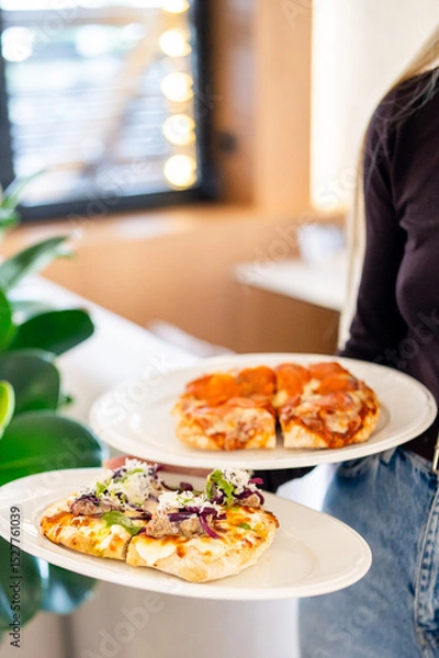Fototapeta A person holds two plates of pizza—one with pepperoni and the other with assorted toppings, including greens and cheese. The blurred background features a window with lights