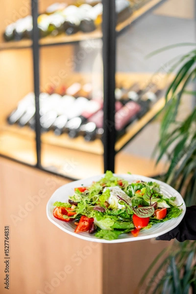 Fototapeta A vibrant salad with fresh vegetables and greens on a white plate, set in a restaurant with a wine rack in the background. Perfect for healthy dining and gourmet presentation.