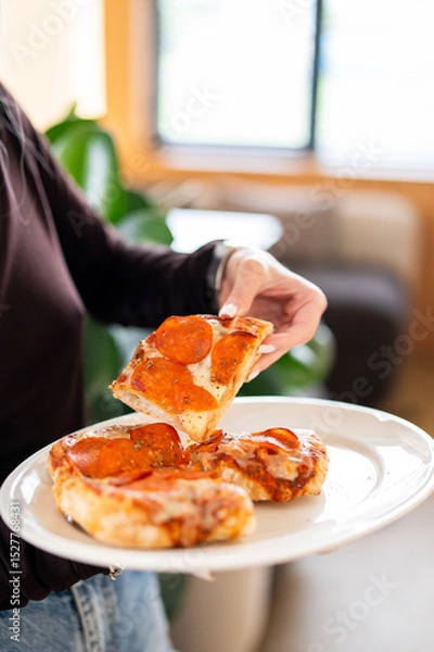 Fototapeta A close-up of a hand lifting a slice of pepperoni pizza from a white plate with two more slices. The melted cheese stretches, enhancing the appeal of this casual dining scene.