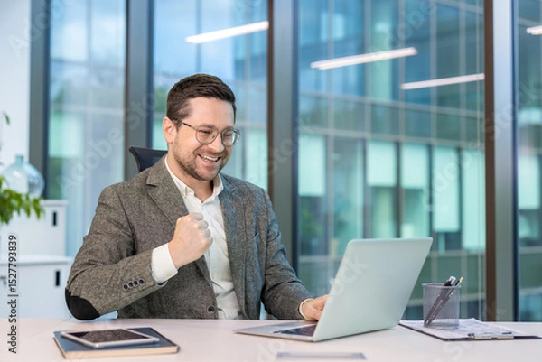 Fototapeta Happy young businessman sitting at a desk in the office, looking at the laptop screen and rejoicing at the news and smiling, showing a victory gesture with his hand