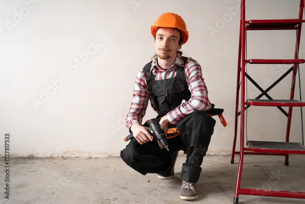 Obraz Portrait of Construction Worker Holding Drill While Sitting Near Ladder at Home Renovation Site
