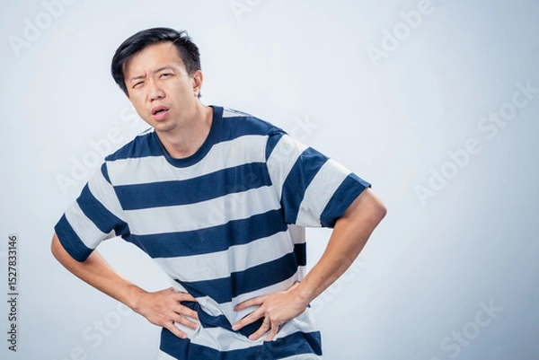 Fototapeta Asian man in striped shirt showing discomfort and fatigue, leaning forward with hands on waist and pained expression, standing against plain studio background