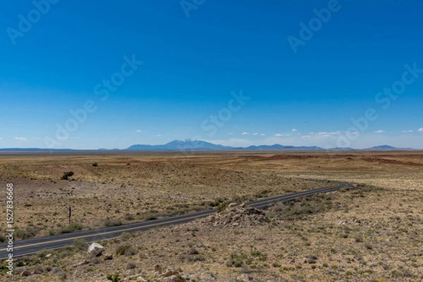 Obraz A winding desert road cuts through the vast, barren landscape near Meteor Crater in Winslow, Arizona, with distant mountains under a clear blue sky.