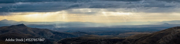 Fototapeta Rolling hills and distant mountains lie under a stormy sky. Iluminating flat-topped rock stands out. Neuquen arid landscape, Argentina