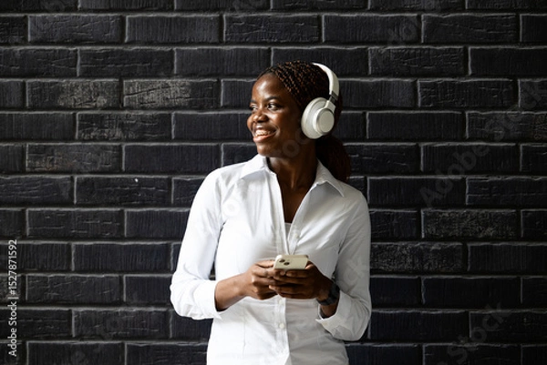 Obraz Portrait of smiling African business woman with smart phone standing by brick wall and looking sideways.