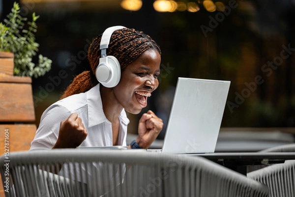 Obraz Close up view of young African woman sitting in cafe and working on laptop computer. Holding fists and celebrating success.