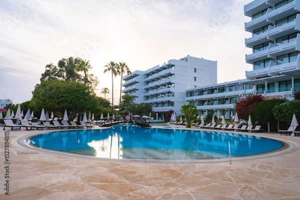 Obraz A large circular pool with lounge chairs, umbrellas, and a decorative bridge, surrounded by palm trees and overlooked by modern white hotel buildings.