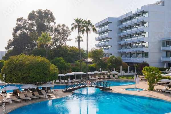 Obraz Modern white resort building with balconies, a large pool, sun loungers, umbrellas, a wooden bridge, lush greenery, and tall palm trees in Cyprus.