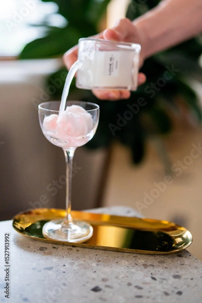 Fototapeta A hand pours a white liquid into a cocktail glass filled with pink cotton candy, placed on a golden tray on a speckled countertop. The scene captures a stylish drink preparation with a luxurious touch