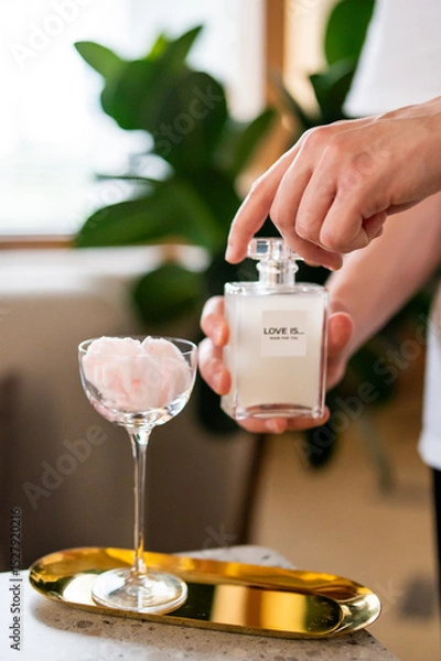 Fototapeta A cotton candy cocktail in a glass, with pink fluffy candy dissolving into the drink. A hand sprays perfume labeled "LOVE IS... MADE FOR YOU" over it.