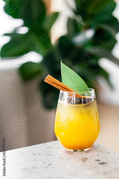 Fototapeta A vibrant yellow-orange cocktail with ice, garnished with a cinnamon stick and a green leaf, placed on a marble surface with a blurred green plant background, evoking freshness and summer vibes